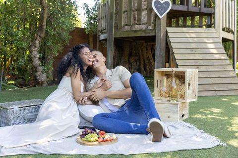 Couple enjoying romantic backyard picnic by treehouse laughing and sharing wine