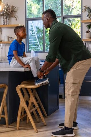 African american father tying sneaker laces for son on kitchen island during morning routine