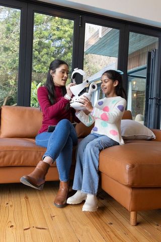 Asian mother and daughter exploring white robot headset on leather sofa in living room