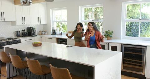 Two women laughing and dancing in modern sunlit kitchen behind marble island, barstools