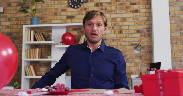 A man with a surprised expression sitting at a table decorated for a birthday celebration with red balloons, gifts, and festive decorations. Bookshelves and a clock adorn the brick wall in the background. Ideal for themes of birthday parties, celebrations, surprises, and cheerful moments at home.