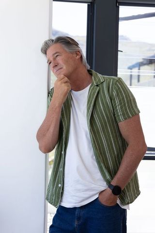 Thoughtful senior man leaning by window and gazing outside in modern sunlit interior