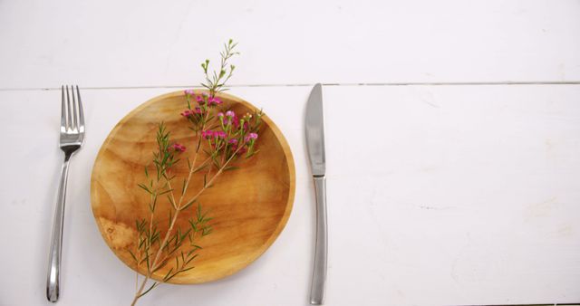 A wooden plate with a sprig of flowers is placed between a fork and knife on a white table, with copy space. It suggests a concept of natural eating or decoration for a dining setting.