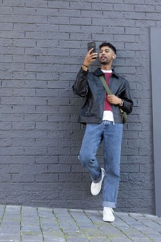 Young man leaning on gray brick wall checking phone while wearing leather jacket and crossbody bag