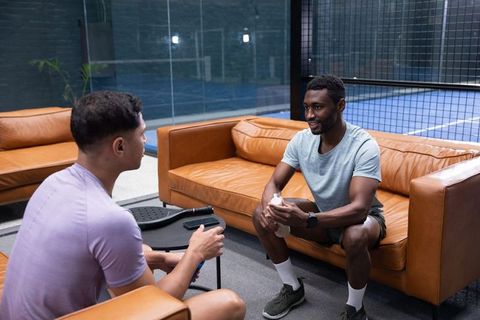 Multiracial men relaxing and chatting on leather sofas after paddle court match