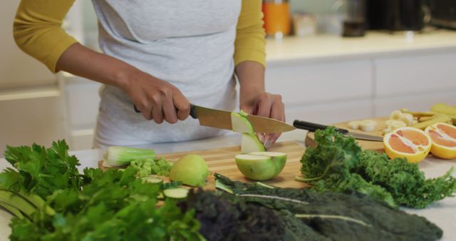 A person slicing fresh vegetables and fruit with a knife in a modern, bright kitchen. The image showcases ingredients like apples, celery, and leafy greens, suggesting a focus on healthy eating and meal preparation. Useful for illustrating content related to nutrition, cooking recipes, healthy lifestyle promotions, or food blogs.
