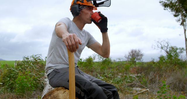 Hardworking lumberjack equipped with safety gear takes a break while drinking coffee. Ideal for use in articles about forestry, physical labor, outdoor professions, and the importance of breaks and hydration. Can also be used in promotional materials for coffee or safety equipment.