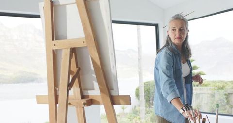 Mature female artist painting by window with scenic lake and mountain view