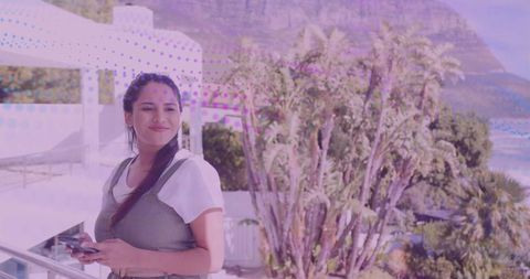Smiling Indian woman leaning on balcony holding smartphone overlooking palm trees and ocean