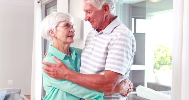 Senior man in striped shirt embracing senior woman in glasses, showing love and happiness at home. Ideal for use in healthcare, retirement living, and lifestyle themes involving seniors and family.