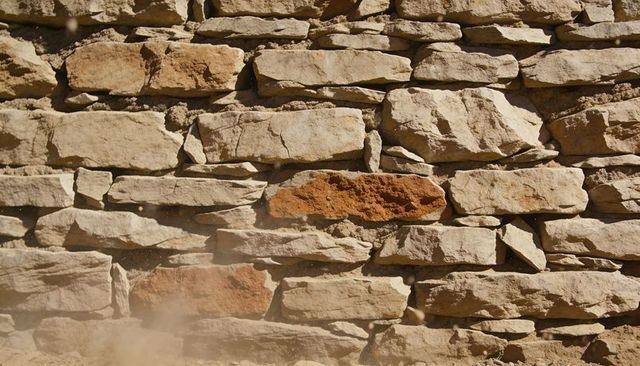Sunlit dry-stacked sandstone wall showing weathered texture, dust haze, reddish stones