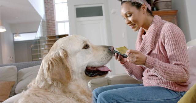 Happy biracial woman with golden retriever dog using smartphone and credit card at home. Lifestyle, animal, communication and domestic life, unaltered.