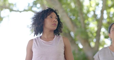 Confident curly-haired woman standing under sunlit tree canopy with friend, outdoor lifestyle