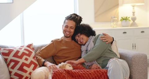 Diverse couple cuddling small white dog on cozy gray sofa, smiling, intimate home lifestyle
