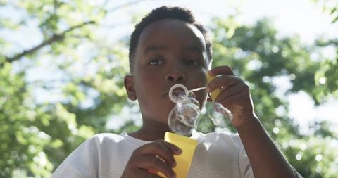 Sunlit boy blowing bubbles with yellow wand in leafy park playful summer childhood moment