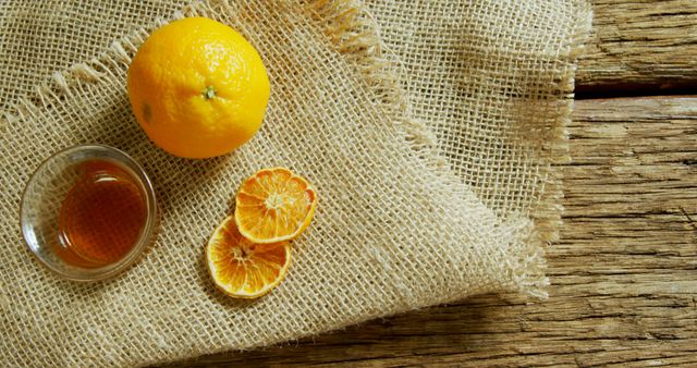 Perfect for use in food blogs, organic health websites, or rustic kitchen aesthetics. Composition showcases a fresh orange with dried slices and a small bowl of honey on a textured burlap and wooden background, emphasizing natural and healthy living concepts.