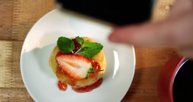 Top view of a gourmet dessert garnished with fresh strawberries and mint on a white plate being photographed by someone. Ideal for food blogs, culinary magazines, restaurant menus, social media campaigns, and photography workshops.