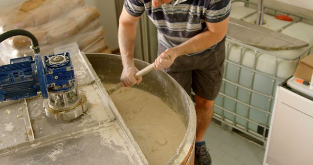 Worker mixing dough in commercial bakery environment, engaging in the process of food production. Industrial equipment and tools visible. Ideal for topics on food production, commercial baking processes, and the bakery industry.
