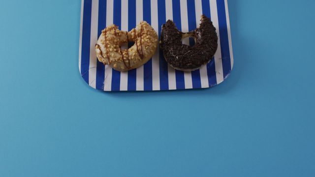 Two donuts with sprinkles placed on a striped plate against blue background. Ideal for marketing dessert products, bakery promotions, or social media posts about indulgent food treats.