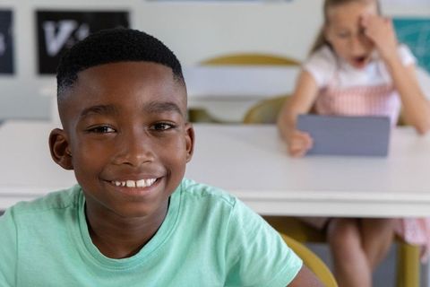 African american boy smiling at desk while classmate using tablet in classroom for education