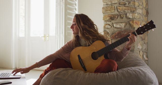 Caucasian woman sitting on beanbag playing acoustic guitar using laptop in sunny cottage living room. simple living in an off the grid rural home.