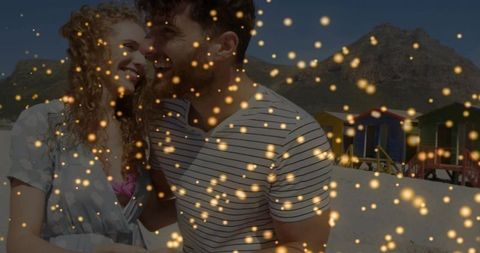 Laughing couple holding hands on sunny beach with colorful huts and golden bokeh glow