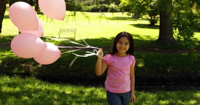 A girl is holding pink balloons and smiling while standing in a sunlit park. Use this image for materials emphasizing childhood joy, outdoor activities, summertime fun, cheerful themes, and promotional content related to parks or events.