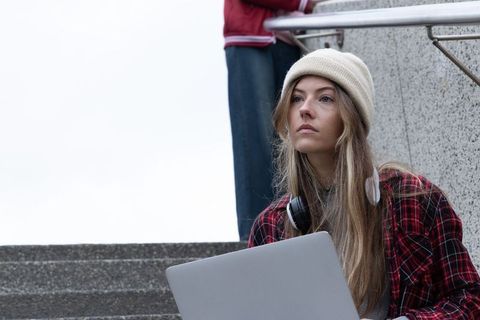 Thoughtful young woman using laptop on urban concrete steps wearing beanie and headphones