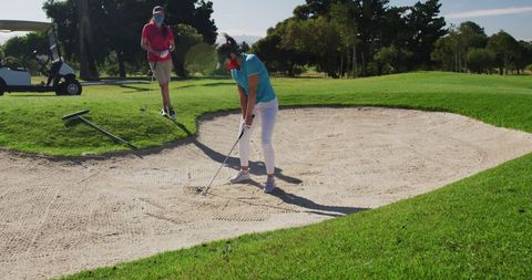 two women play golf while wearing face masks on a sunny day, focusing on a shot from a bunker. the image captures leisure activities and outdoor sports continuing amid health precautions. ideal for topics on pandemic lifestyle, health-conscious sports, and recreation with safety measures. useful for sports magazines and health safety articles.