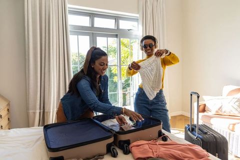 Friends packing for vacation laughing while folding beaded top in sunlit bedroom