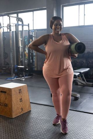 African american woman carrying yoga mat in gym wearing mauve activewear and smiling