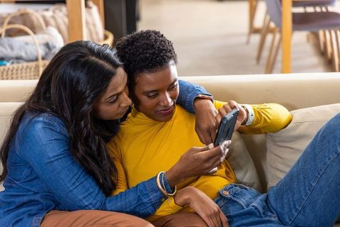 Multiethnic female couple cuddling on sofa, sharing smartphone, cozy intimate moment