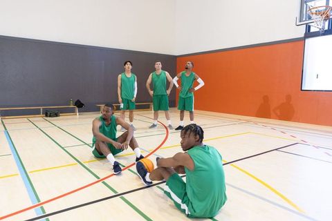 Diverse basketball team resting and preparing on indoor court wearing green uniforms