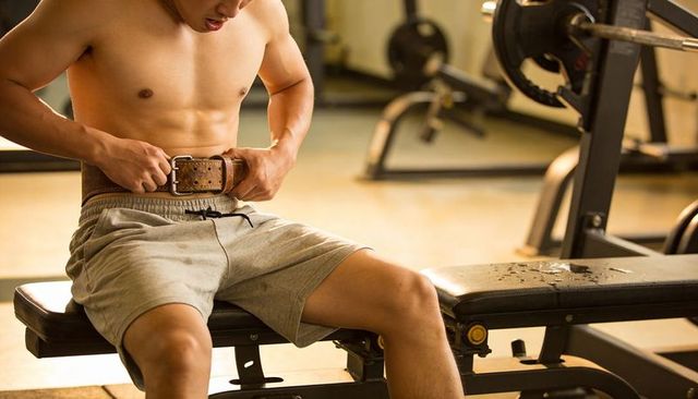 Shirtless asian man fastening leather weightlifting belt on gym bench preparing heavy lift