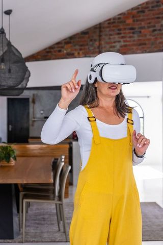 Mature woman wearing white vr headset and yellow overalls exploring virtual home