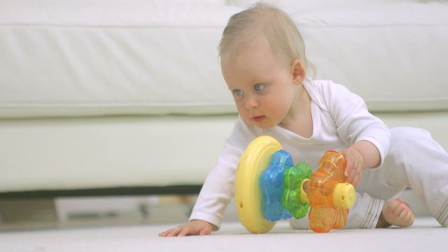 Baby wearing white outfit plays with colorful plastic toys on floor in well-lit indoor setting. Ideal for parenting, childcare, early childhood development, family lifestyles, toy advertisements, and home interiors.