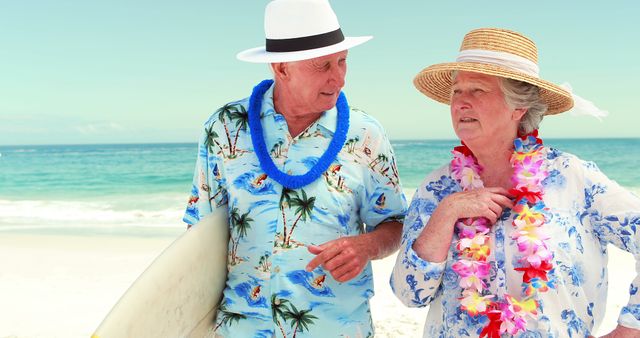 Elderly couple wearing Hawaiian shirts and leis, standing on beach. Woman in straw hat holding lei, man holding surfboard. Perfect for vacation, travel, retirement, senior living, summer enjoyment concepts.