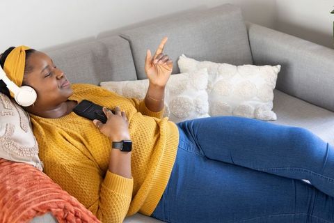 Relaxing african american woman listening to music on couch wearing mustard sweater