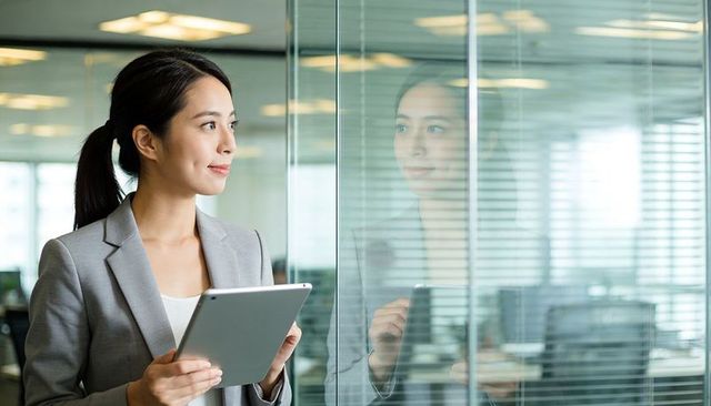 Asian businesswoman holding tablet and looking through glass in modern corporate office
