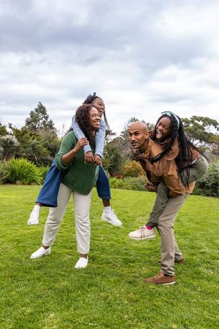 African american family laughing in park parents giving piggyback rides on grassy lawn