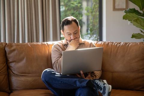 Mature asian man working on laptop on leather sofa, thinking during remote work
