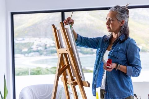 Senior woman painting by large window with coastal view in bright home studio