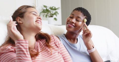 Multiracial friends sharing earbuds, laughing and relaxing on sunlit white couch