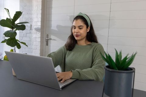 Young woman working on laptop in minimalist home office with plants and green sweater