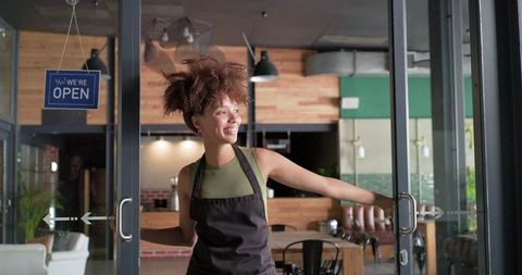 Smiling african american barista opening cafe door with 'open' sign welcoming customers