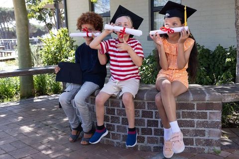 Diverse children celebrating graduation wearing mortarboards holding diplomas outdoors