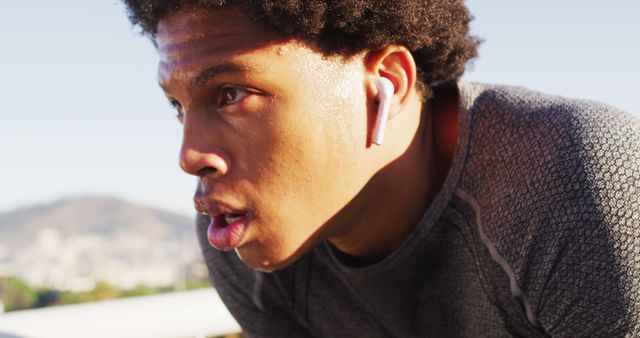 Young athlete seen taking a moment to breathe during an intense outdoor workout. Notice the sweat on his face and wireless earphones, emphasizing dedication and modern fitness trends. Perfect for illustrating themes of hard work, fitness goals, sports achievements, and motivation in advertising, health-related articles, and fitness blogs.