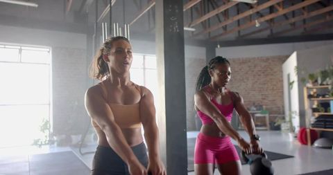 Swinging kettlebells two athletic women performing strength workout in sunlit studio