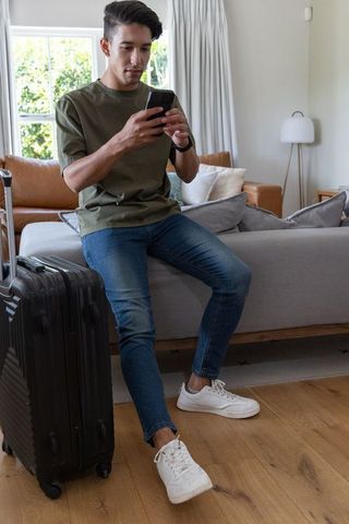 African american man checking phone on sofa with suitcase in modern living room