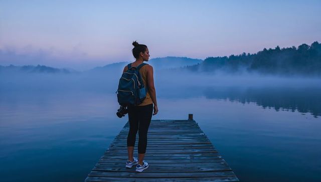 Standing woman on wooden dock at misty dawn lake carrying backpack and camera